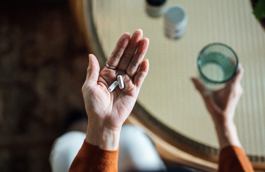 Photo looking down to a woman's hands, one holding a glass of water, in the other hand, some pills