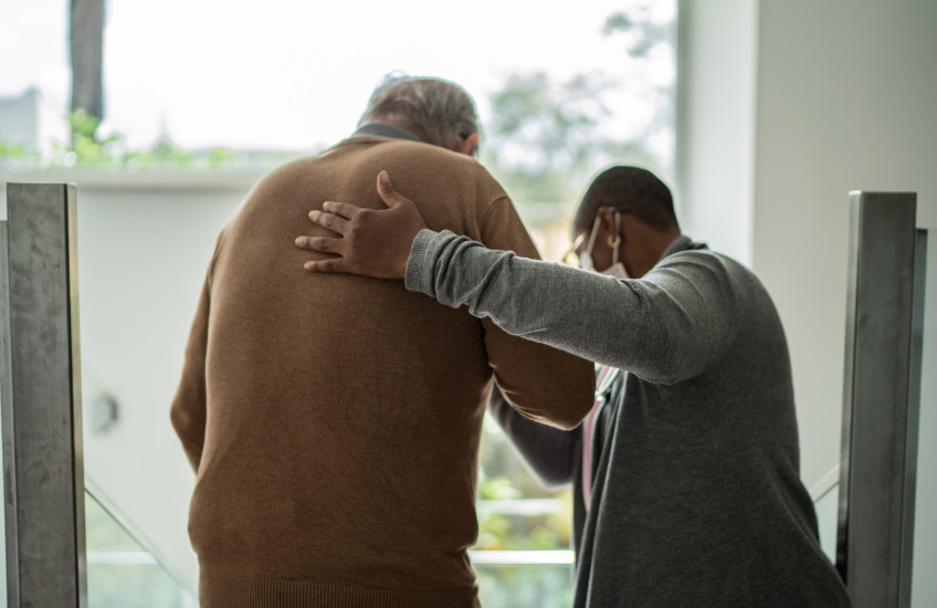 Rear view photo of health care worker helping a patient to walk, inside a room with windows