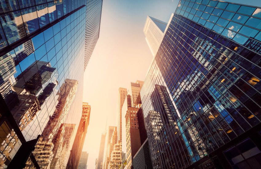 Photograph of tall office buildings, taken from the street, looking up to a sunny sky and tops of skyscrapers