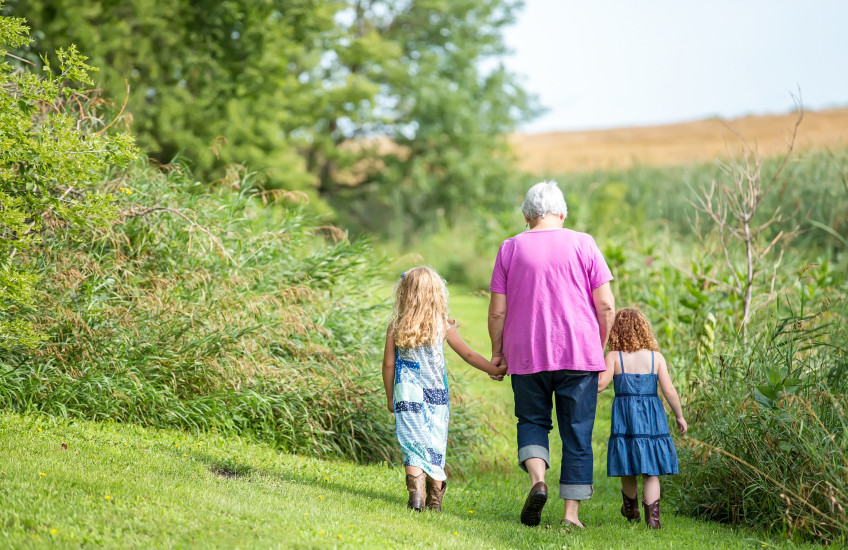 Woman in pink top walking, holding hands with two little girls, backs to camera, in a garden