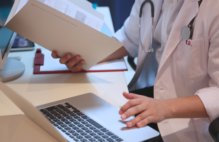 Photo of physician holding a pen and a notebook and typing on a computer keyboard