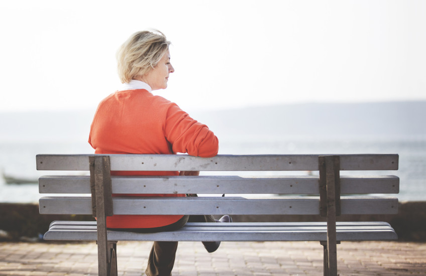 Woman with light hair and orange sweater sits alone on a bench overlooking water