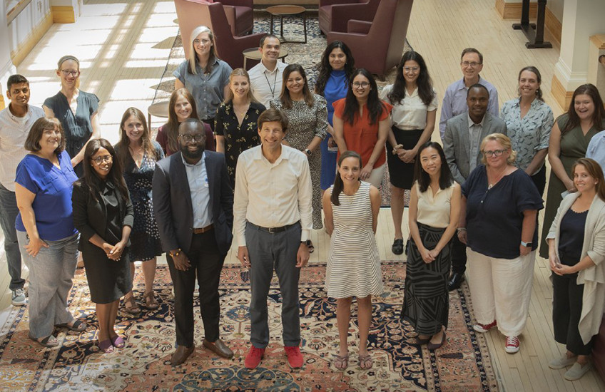 Photographed from above, 24 people stand in the center of a sunlit room smiling up at the camera.