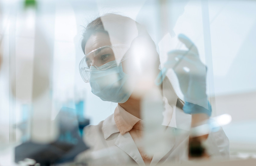 A woman in goggles mask and lab coat working at a laboratory table seen through the reflections and refractions on a safety glass window.