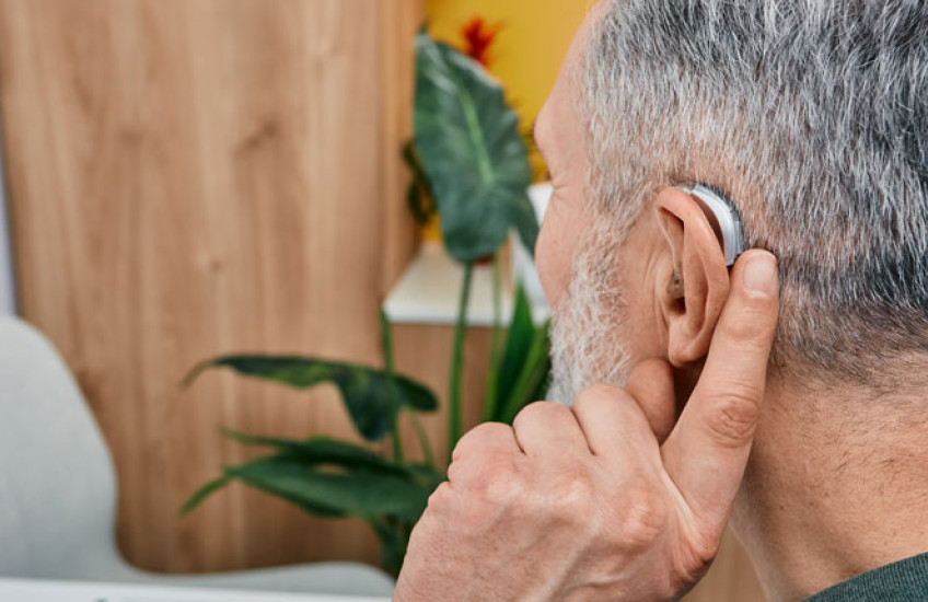 back of the head of a white grey-haired man holding a finger to his ear that has a hearing aid