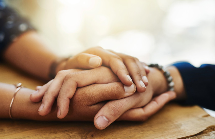 Closeup of two people’s hands clasped together on a table in a comforting manner.