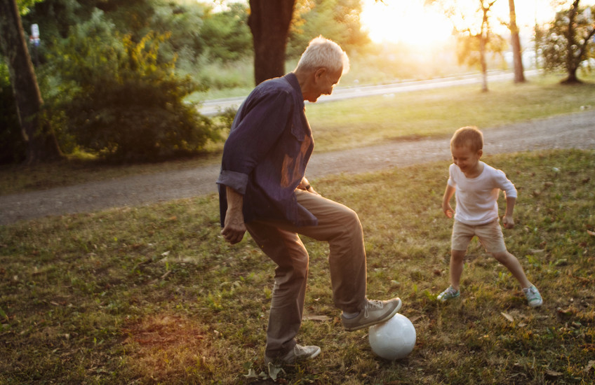 Caught in the middle of a one-on-one soccer game, a white-haired man stands with his foot on a soccer ball, a small child faces him, poised to take the ball.