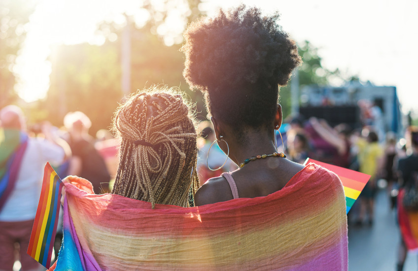 Rear view image of two people with braided, long hair, wrapped in a rainbow scarf walking with other people waving rainbow flags 