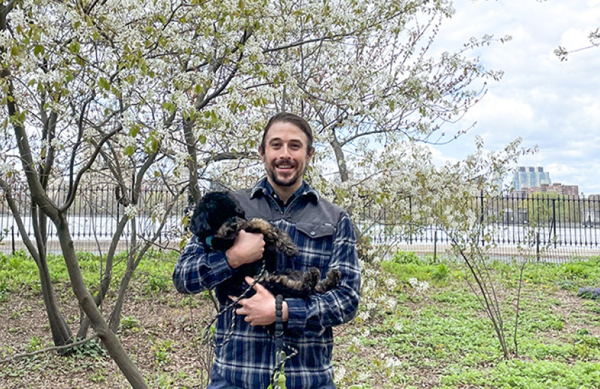 Nelson LaMarche with his miniature poodle in a NY city park with blooming pear tree and Hudson River behind him