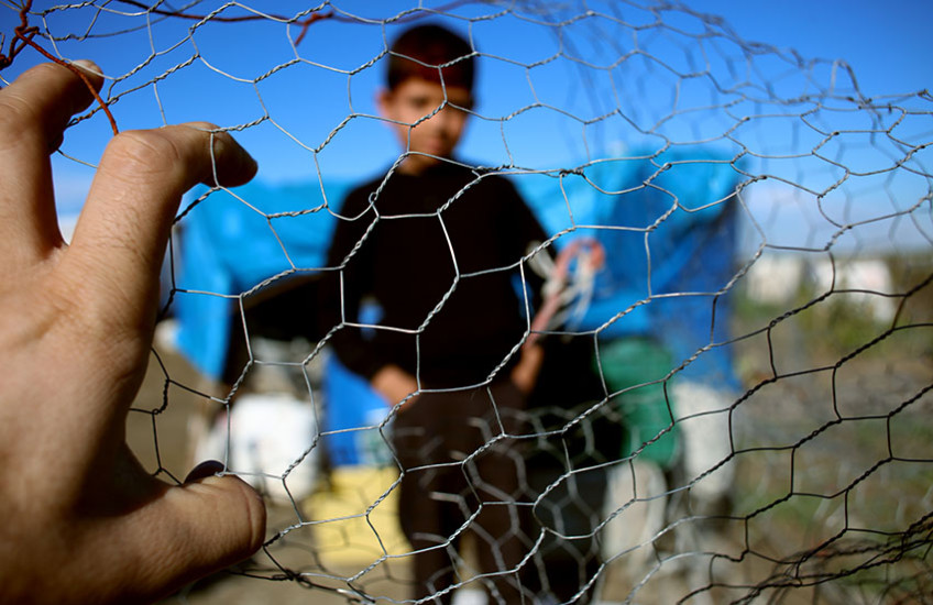 A stock photo of a hand holding a fence with a blurred image of a boy on the other side of the fence.
