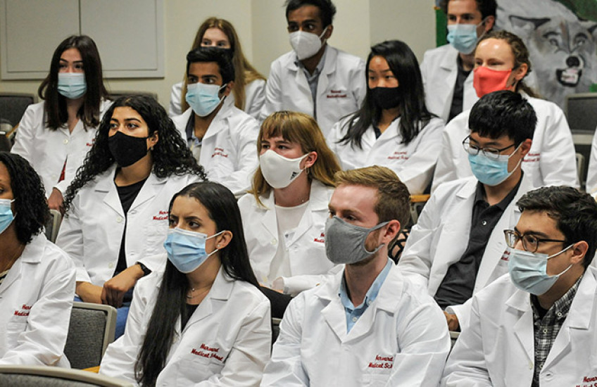 17 students wearing masks and in white coats that say Harvard Medical School seated in auditorium seats