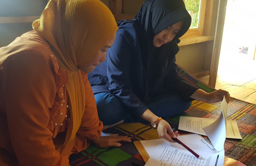 Two women, one in orange and one in blue seated on a colorful rug look at documents together.