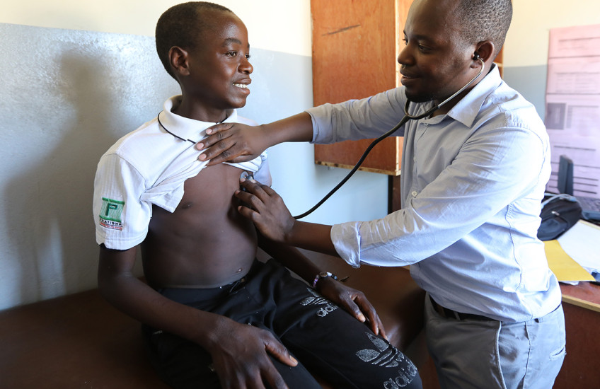 A teenage boy sits on an examination table in a clinic as a man listens to his heart with a stethoscope.