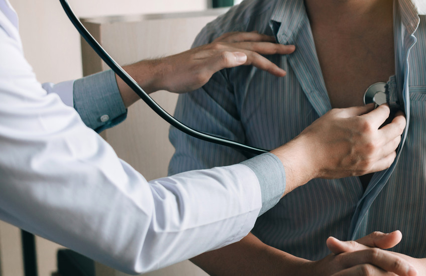 A person in a white jacket is using a stethoscope to listen to the heartbeat of person with a loosely buttoned striped shirt.