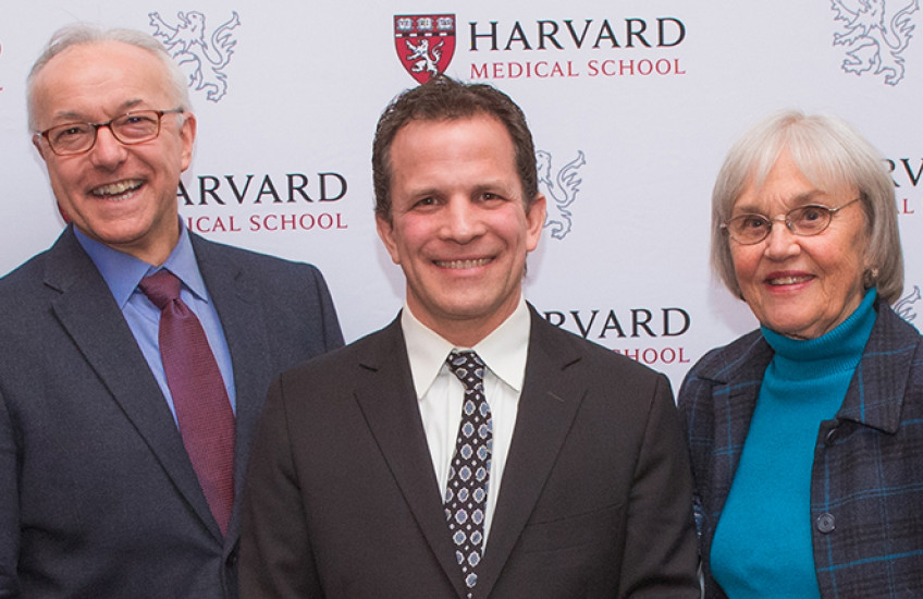 Two men and a woman standing together smiling in front of a banner with HMS logo