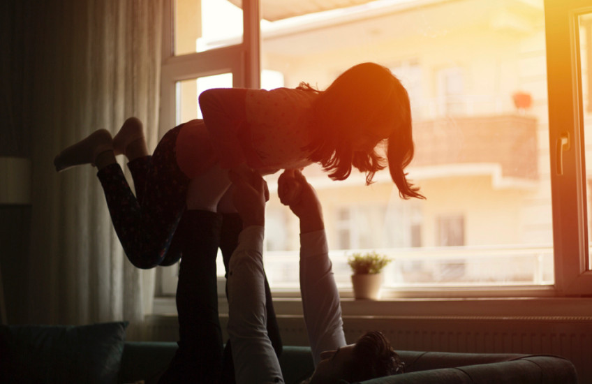 A father plays with his daughter backlit by sunset, seen through the window.