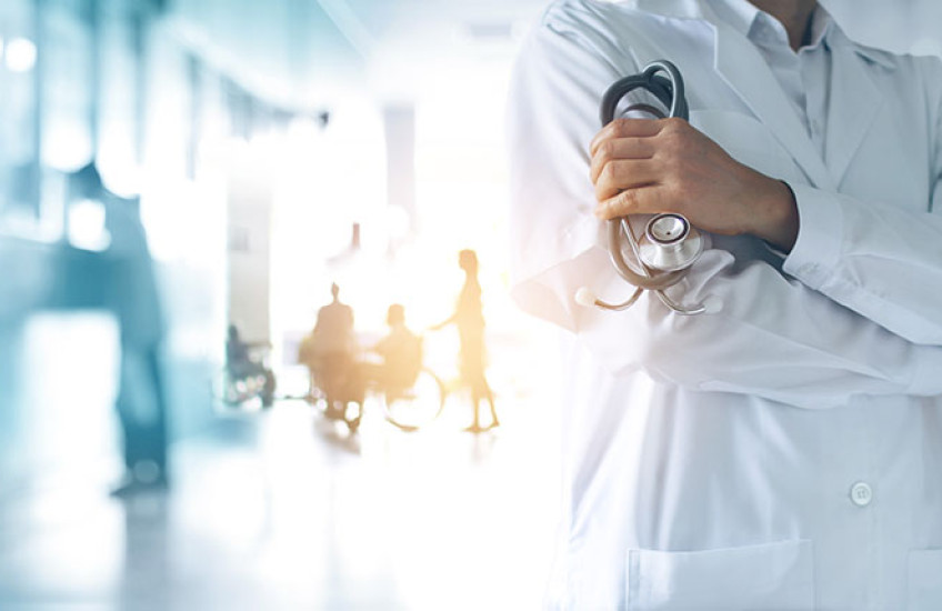 A physician with arms crossed stands in front of a blurry, overexposed background of a mostly empty waiting room.