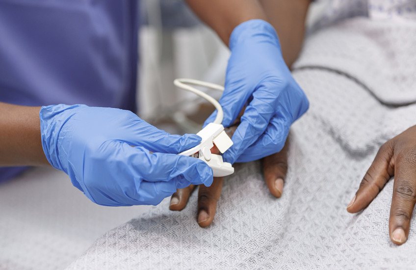 Close up shot of a medical professional placing a pulse oximeter on the finger of a Black hospitalized patient who is lying in bed.