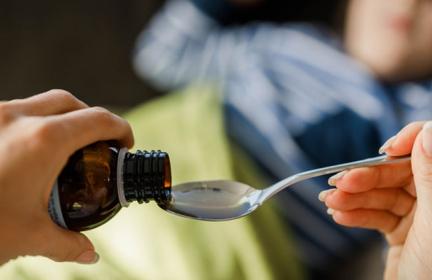 Closeup of adult women's hands pouring cough medicine into a spoon with child blurred out in background
