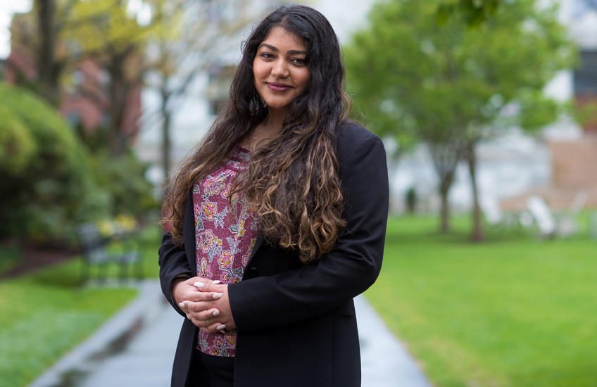 A woman with long, curly hair wearing a dark blazer and a flower-patterned shirt stands on a path between spring green bushes and lawn, an out of focus marble building in the background.