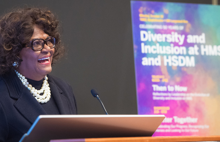 Alt Text:  A woman standing at a lectern, speaking in front of a colorful, out-of-focus poster