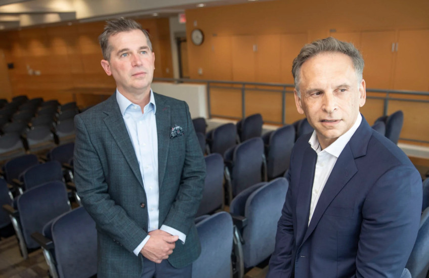 Two men in suits lean against chairs in an auditorium. 