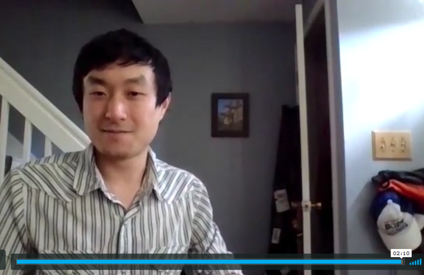 Screen shot of young Asian-American man with bannister and framed photo on wall in background