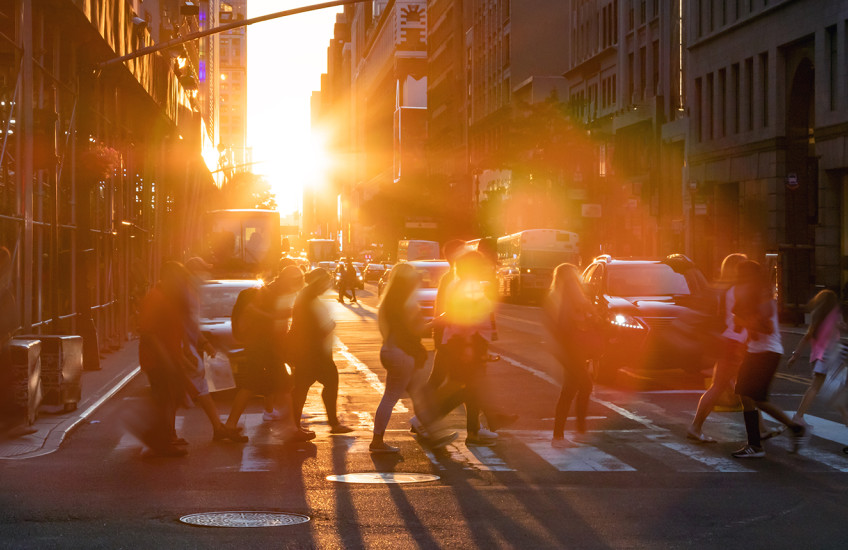 A group of people cross a street at a crosswalk, backlit by the glow of the sun and surrounded by a halo of rainbowed lens flare.
