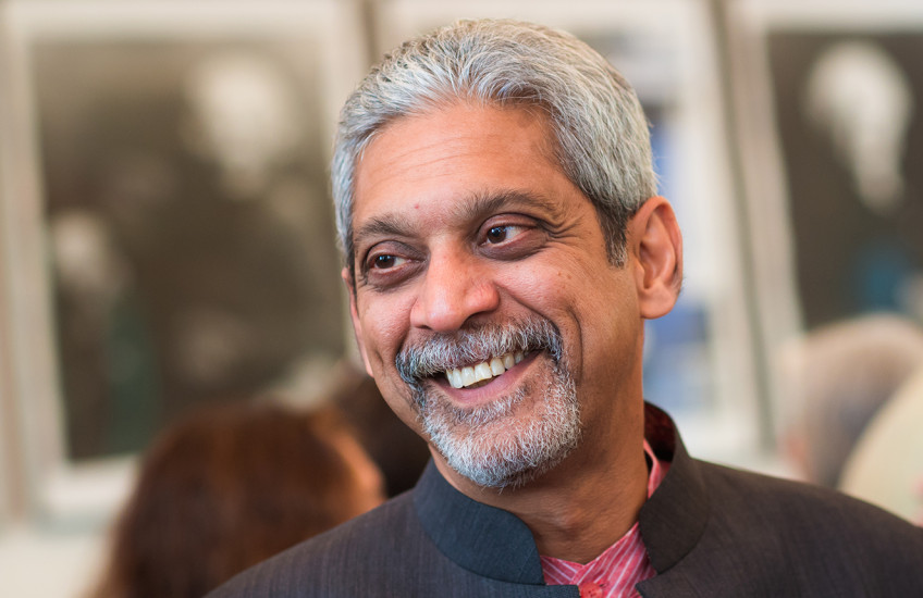 A silver-haired man, wearing a black jacket and a red shirt, smiles at someone off camera.
