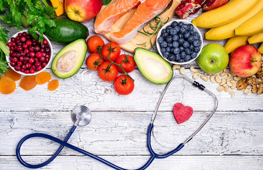 A stock photo of fruits and vegetables on a table next to a stethoscope with a red heart between the stethoscope earpieces.