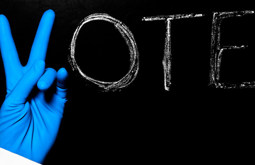 Photo of chalkboard with the word vote - the V is being made by the hand of a person with surgical gloves on.