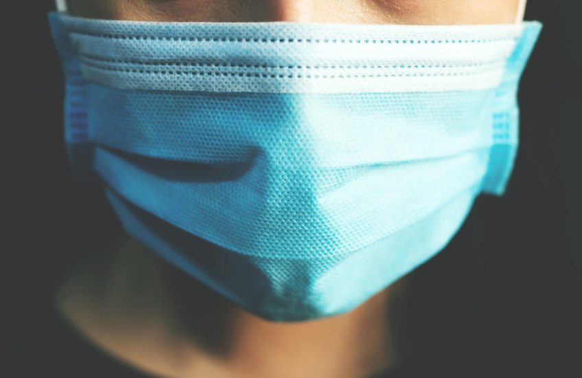 Closeup of the face of a person wearing a paper medical mask in front of a dark background.