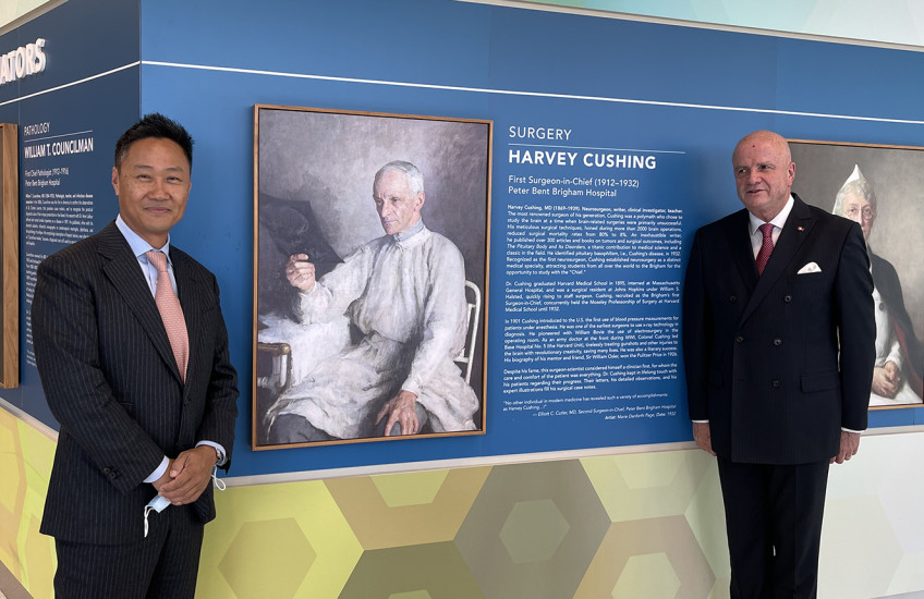 In a hallway painted with colorful geometric patterns, two men in suits stand on either side of a portrait of a man in surgical gowns reviewing an X-ray of a patient’s skull. The portrait is labeled “Harvey Cushing.”