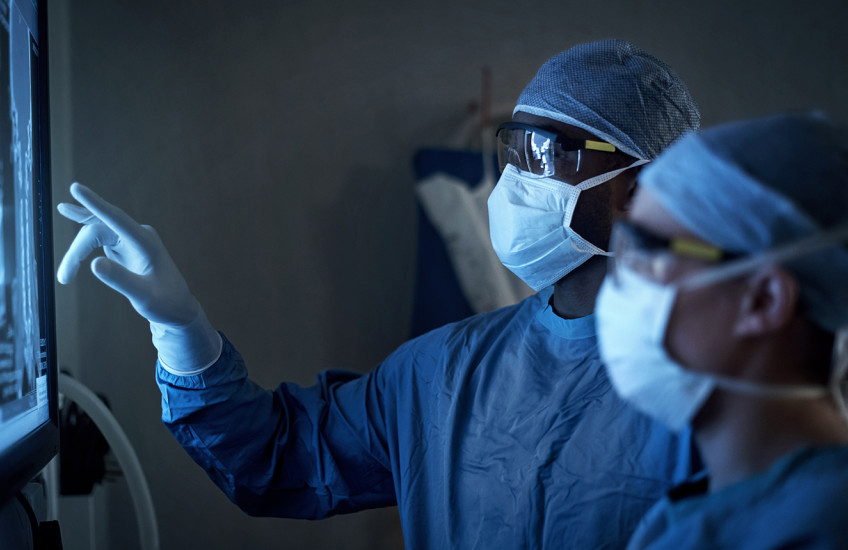 Two figures wearing surgical masks, gowns, and caps examine a backlit X-ray image