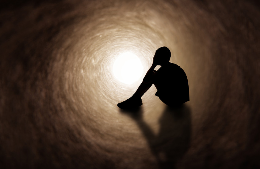 The silhouette of a young person in shorts and t-shirt covering their face as they sit in a dark tunnel.