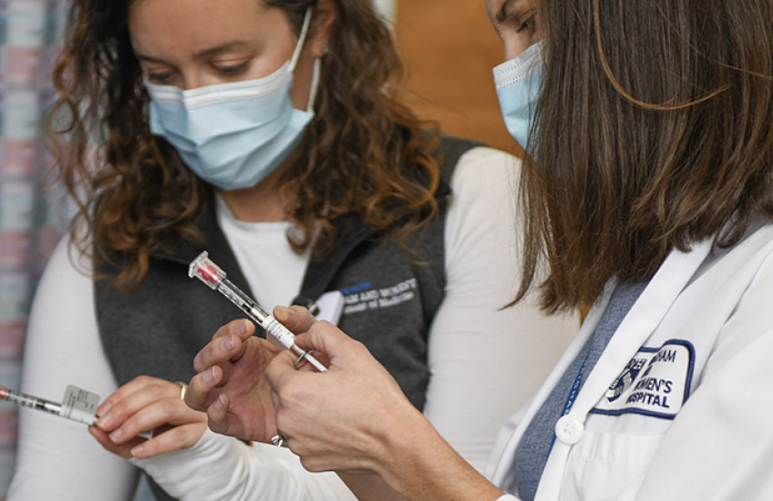 Two women in white coats and surgical masks hold syringes labeled "COVID-19 vaccine"