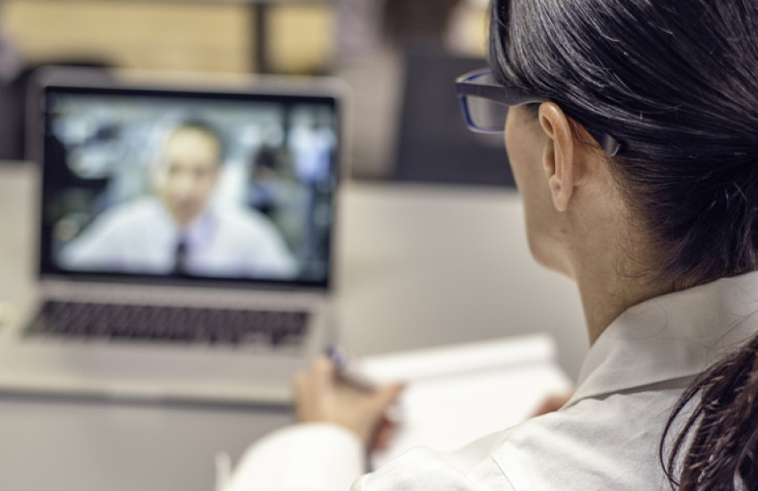 Looking over a doctor's shoulder, we see a patient video conferencing on the doctor's laptop.