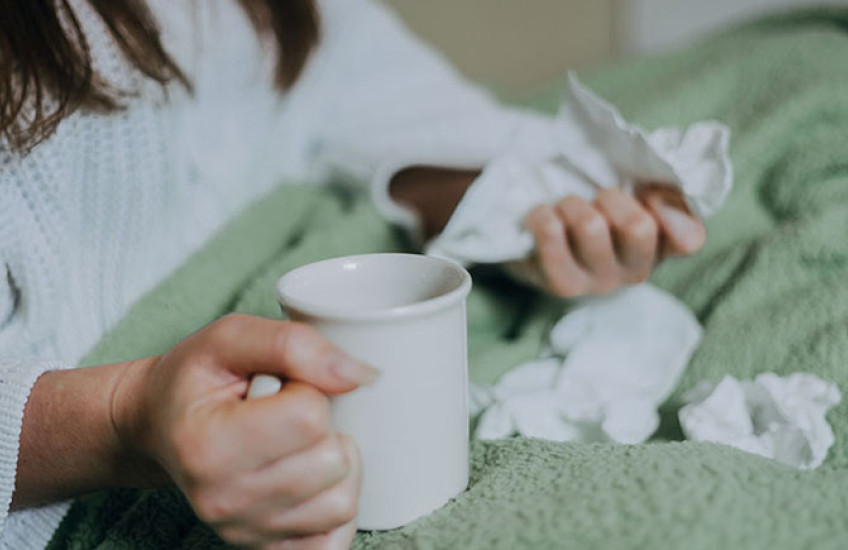 a person sitting in bed under a blanket, holding tea and surrounded by crumpled facial tissues