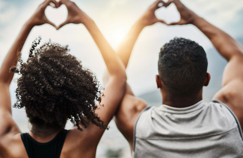 Photo of a Black young man and young woman making heart signs with their hands, backs facing the camera