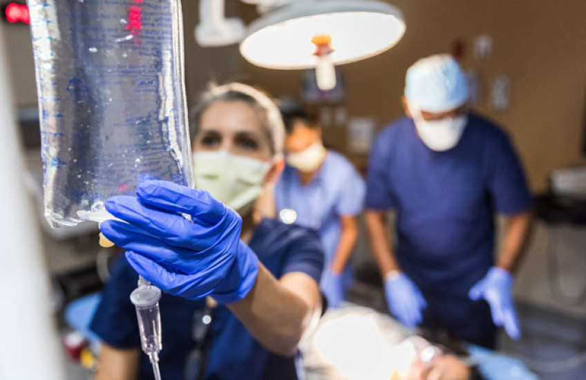 Out-of-focus photo of ER nurse checking an IV bag with a surgeon looking at a patient on a gurney behind her