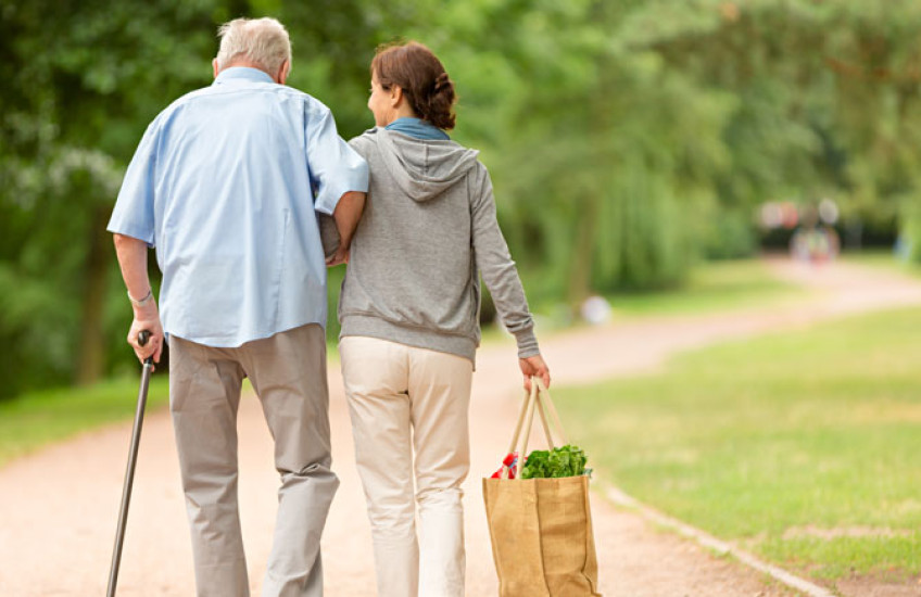 Back view of a woman walking with an older man supported by a cane along a dirt path with trees and grass surrounding