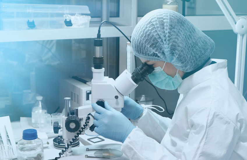Photo of a female scientist in white lab coat peering into a microscope at a lab bench