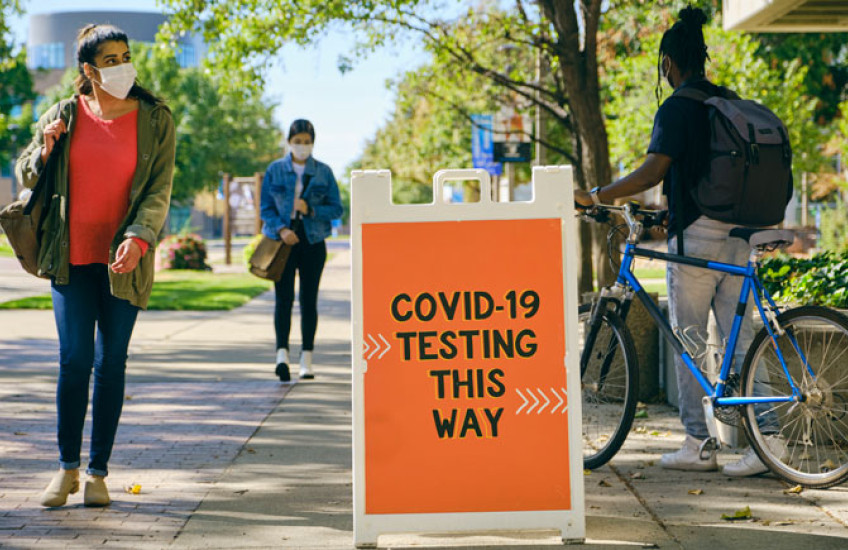 An orange sign that says "COVID-19 testing this way" on a college campus with students walking nearby