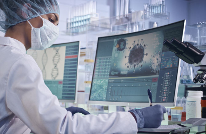 A scientist sits writing notes in front of a computer monitor showing a COVID-19 virus