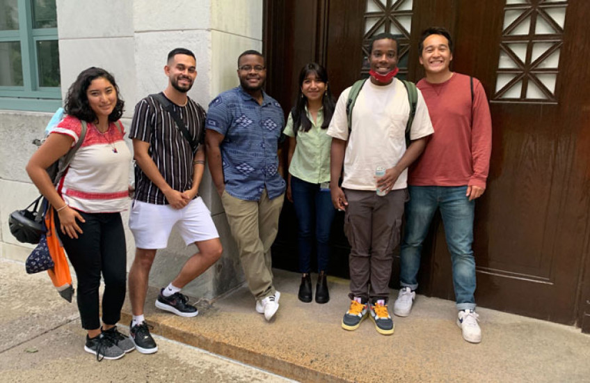Six students pose for a photo against a stone building and a wooden door