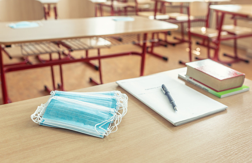 A stack of surgical masks, a pad of paper and pen, and a pile of books sit on a table in an empty classroom