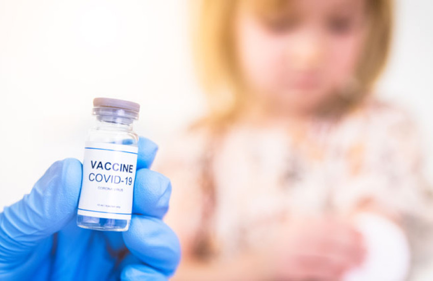 Blue-gloved hand holding COVID vaccine vial and blurry child in background	 