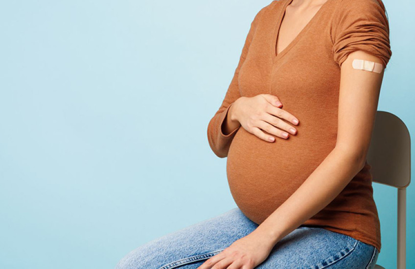 Cropped stock image of a woman with light brown skin sitting in a hair with a hand on her pregnant belly. A bandage on her arm indicates a recent COVID vaccination.