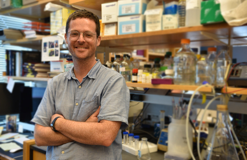 Young white man stands smiling with arms crossed in front of lab bench full of bottles