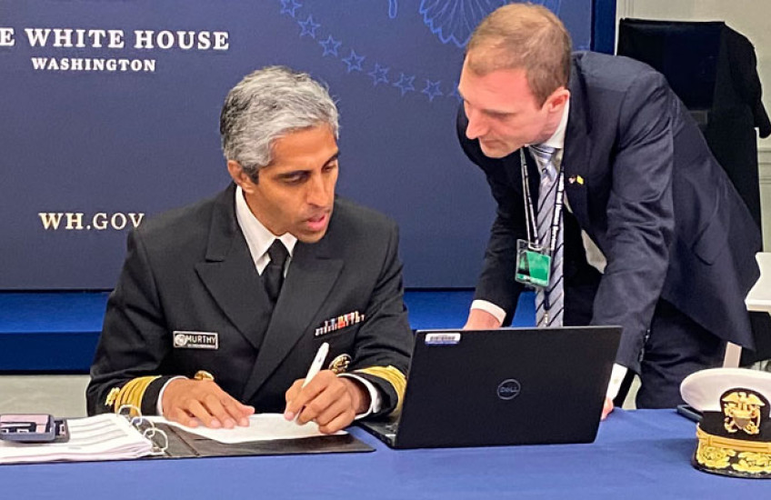 Murthy in uniform seated at table looking at laptop with Beckman to right leaning in; a backdrop behind them reads The White House